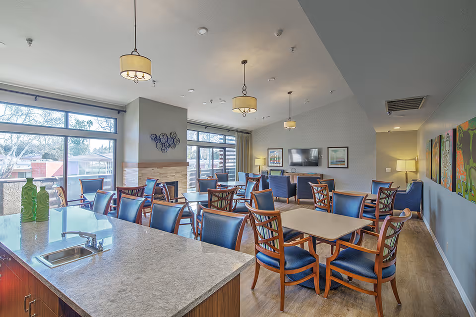 A bright and spacious senior living common area with multiple wooden tables and blue cushioned chairs arranged neatly. Large windows let in natural light, and there is a fireplace with a decorative wall piece above it. The room also features a TV mounted on the wall, colorful artwork, and modern pendant lights hanging from the ceiling. A countertop with a sink is visible in the foreground.