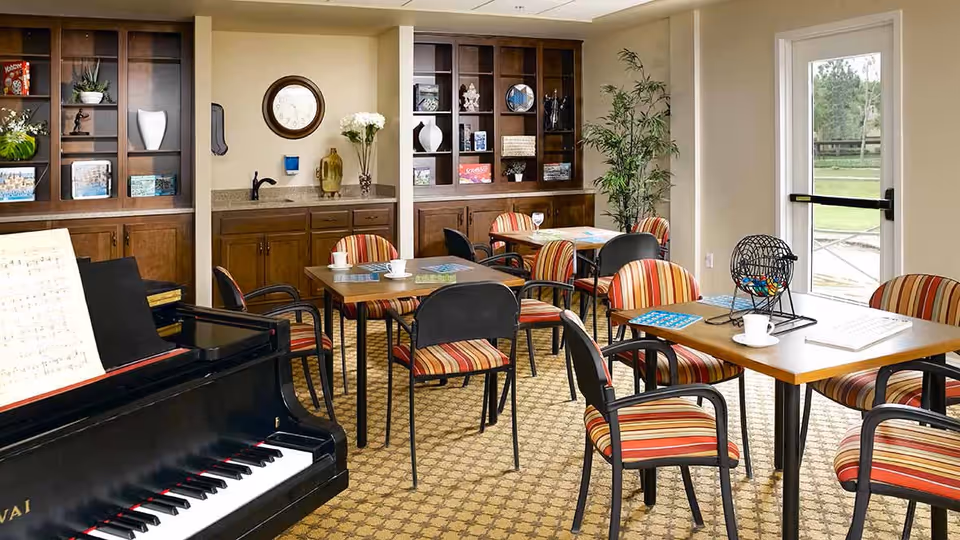 A cozy activity room with several tables and striped chairs arranged for group activities. A black piano with sheet music is visible in the foreground. The room has built-in wooden shelves with decorative items and books, a small sink area, and a door leading outside with a view of greenery.