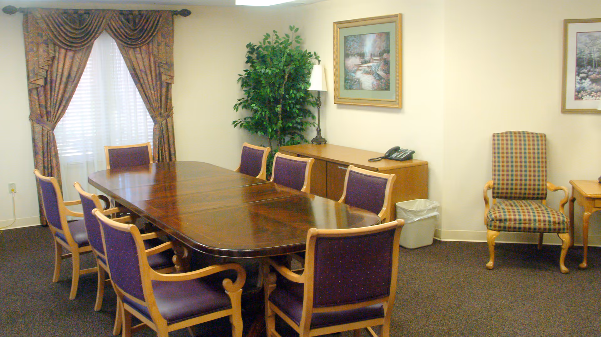 A dining room with a large polished wooden table surrounded by eight purple cushioned chairs with wooden frames. The room has beige walls, a window with patterned curtains, a green potted plant, a wooden sideboard with a telephone and lamp, a trash bin, and framed artwork on the walls. There is also a single armchair with a plaid pattern and wooden legs.