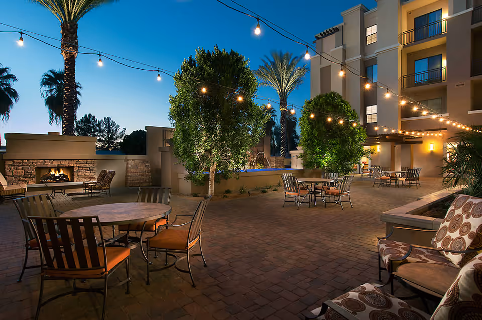 Outdoor patio area at dusk with string lights overhead, several round tables with chairs, cushioned seating, a lit fireplace built into a stone wall, palm trees, and a multi-story building in the background.