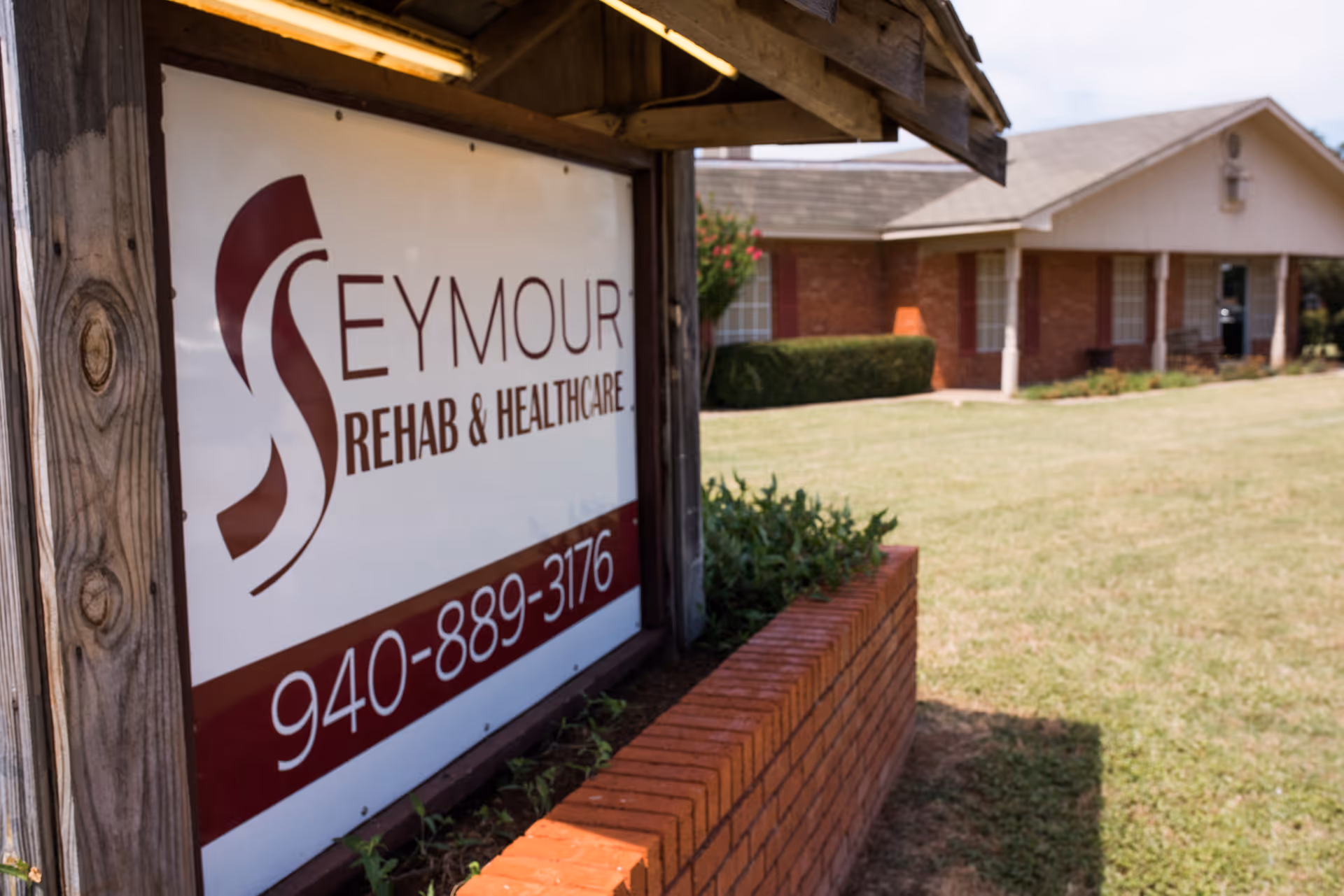 Outdoor view of Seymour Rehabilitation and Healthcare facility sign with a phone number, set in a wooden frame with a brick planter in front, and a brick building with a lawn in the background under a partly cloudy sky.