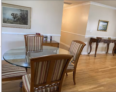 A dining area with a round glass table surrounded by four wooden chairs with striped upholstery. The room has light-colored walls with white trim, hardwood flooring, and two framed paintings on the walls. There is a wooden side table with decorative items in the background.