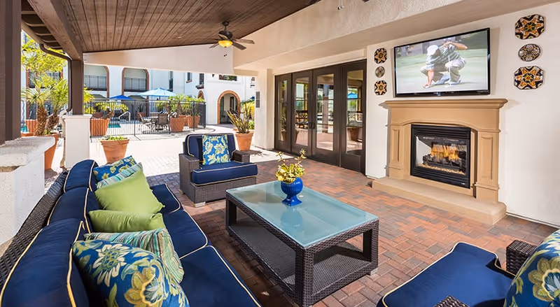 Covered outdoor seating area with blue cushioned sofas, glass coffee table, fireplace and TV overlooking a pool courtyard.