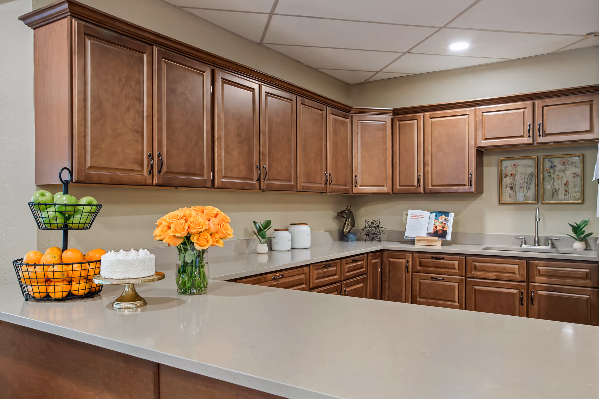A kitchen area with wooden cabinets, a beige countertop, a two-tiered fruit basket with green apples and oranges, a white cake on a gold stand, a vase of orange roses, and decorative items including a cookbook on a stand and framed floral artwork on the wall.