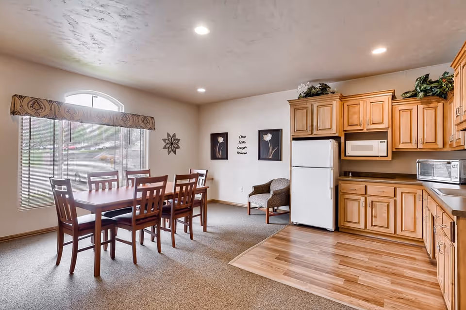 Bright dining area with a wooden table and chairs adjacent to a kitchenette with wooden cabinets and a white refrigerator.
