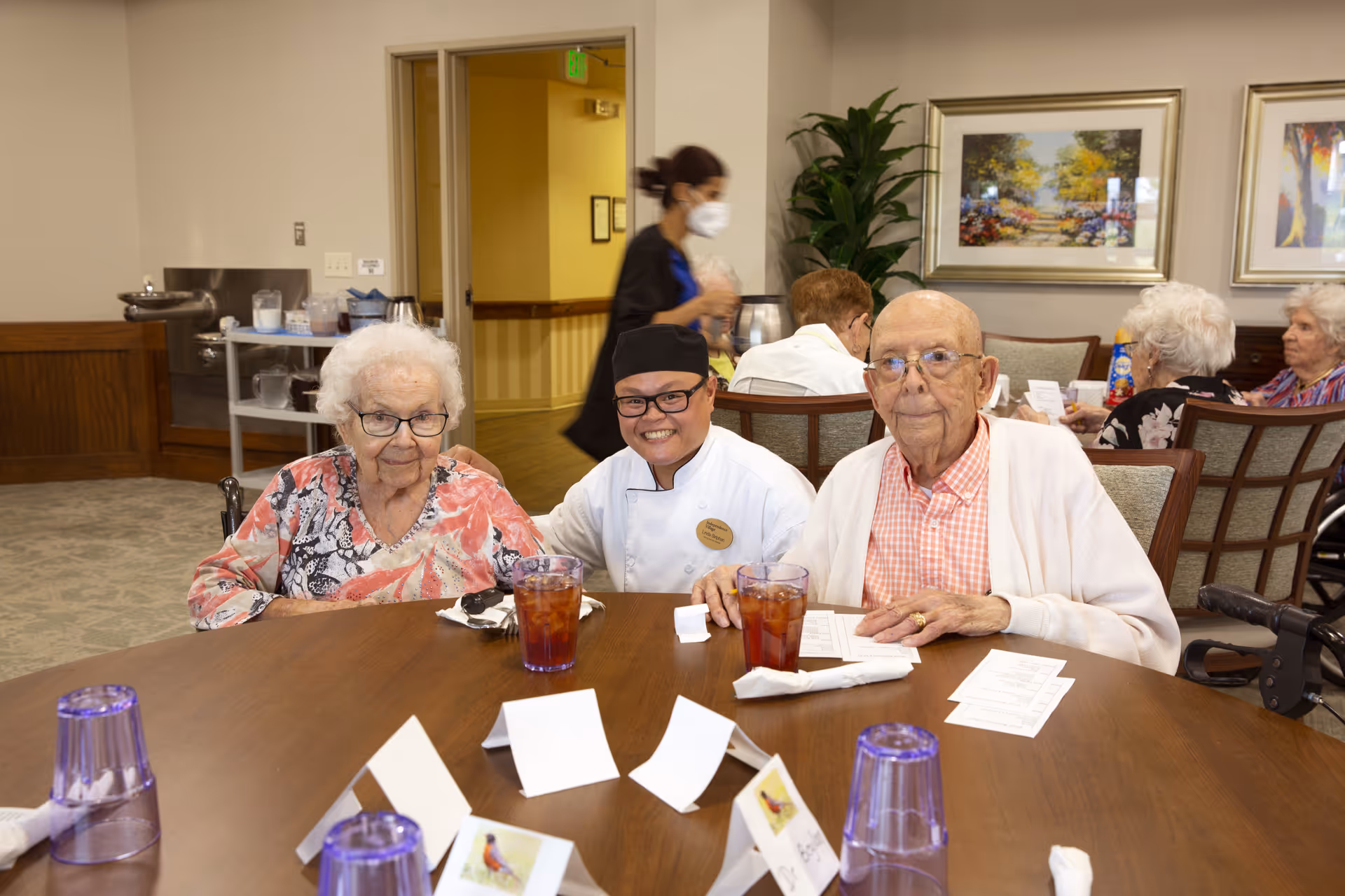 Two elderly individuals sitting at a round dining table with a smiling staff member in a chef uniform between them. The table has glasses of iced tea, napkins, and place cards. Other elderly people are seated at tables in the background, and a staff member wearing a mask is walking by.