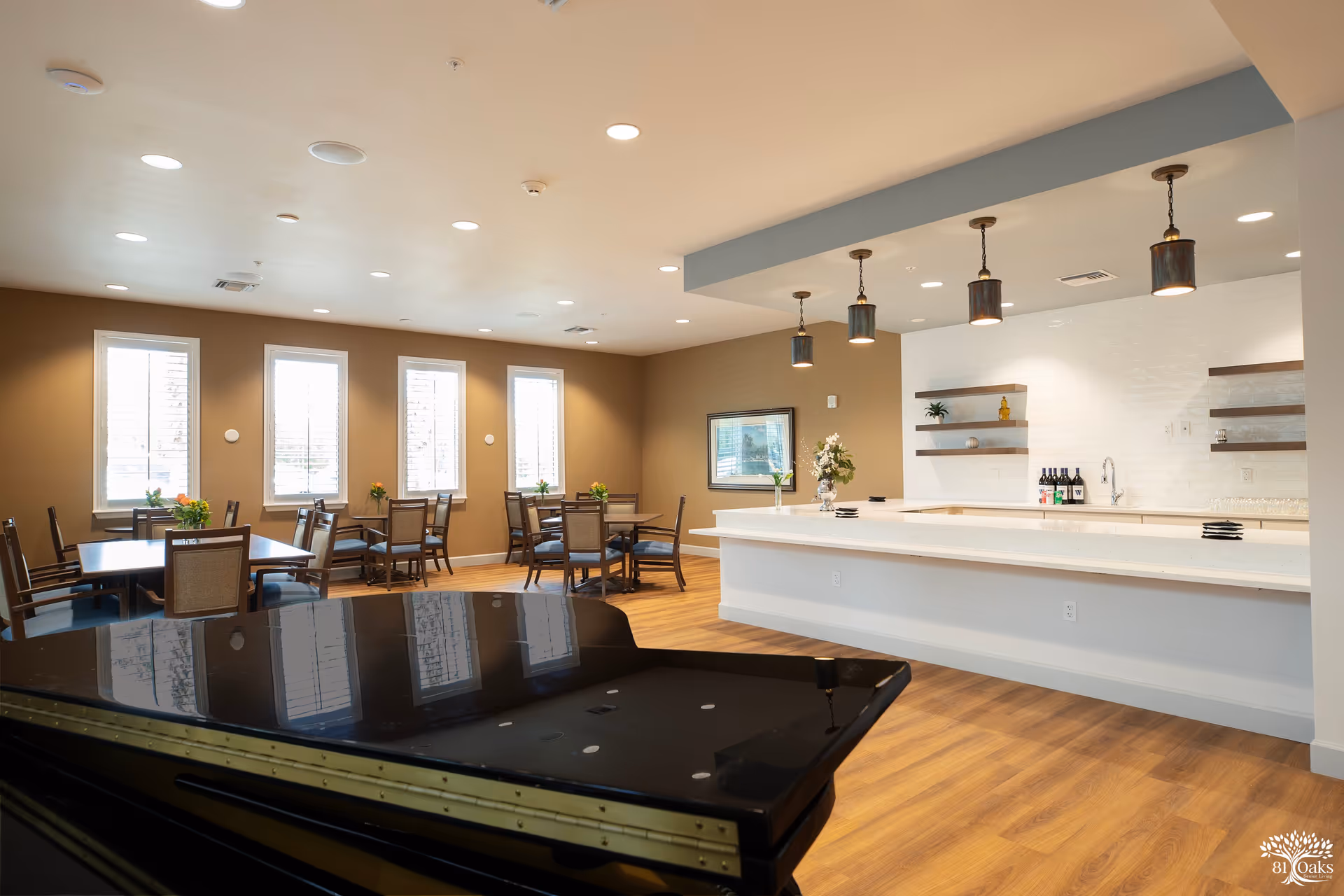 A spacious and well-lit dining area in a senior living facility featuring multiple tables with chairs, a white countertop bar with hanging pendant lights, decorative shelves with plants and bottles, and a black grand piano in the foreground. The room has wood flooring and several windows with white blinds.