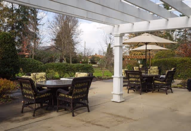 Outdoor patio area with two round tables surrounded by wicker chairs with cushions. One table has a large beige umbrella. The patio is covered partially by a white pergola and surrounded by green bushes and trees under a partly cloudy sky.
