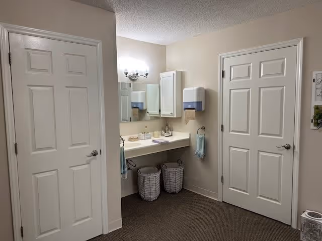 Interior view of a room with two closed white doors, a wall-mounted sink with a mirror above it, two laundry baskets underneath, a wall-mounted paper towel dispenser, and a towel ring with a blue towel. The walls are beige and the floor is carpeted.
