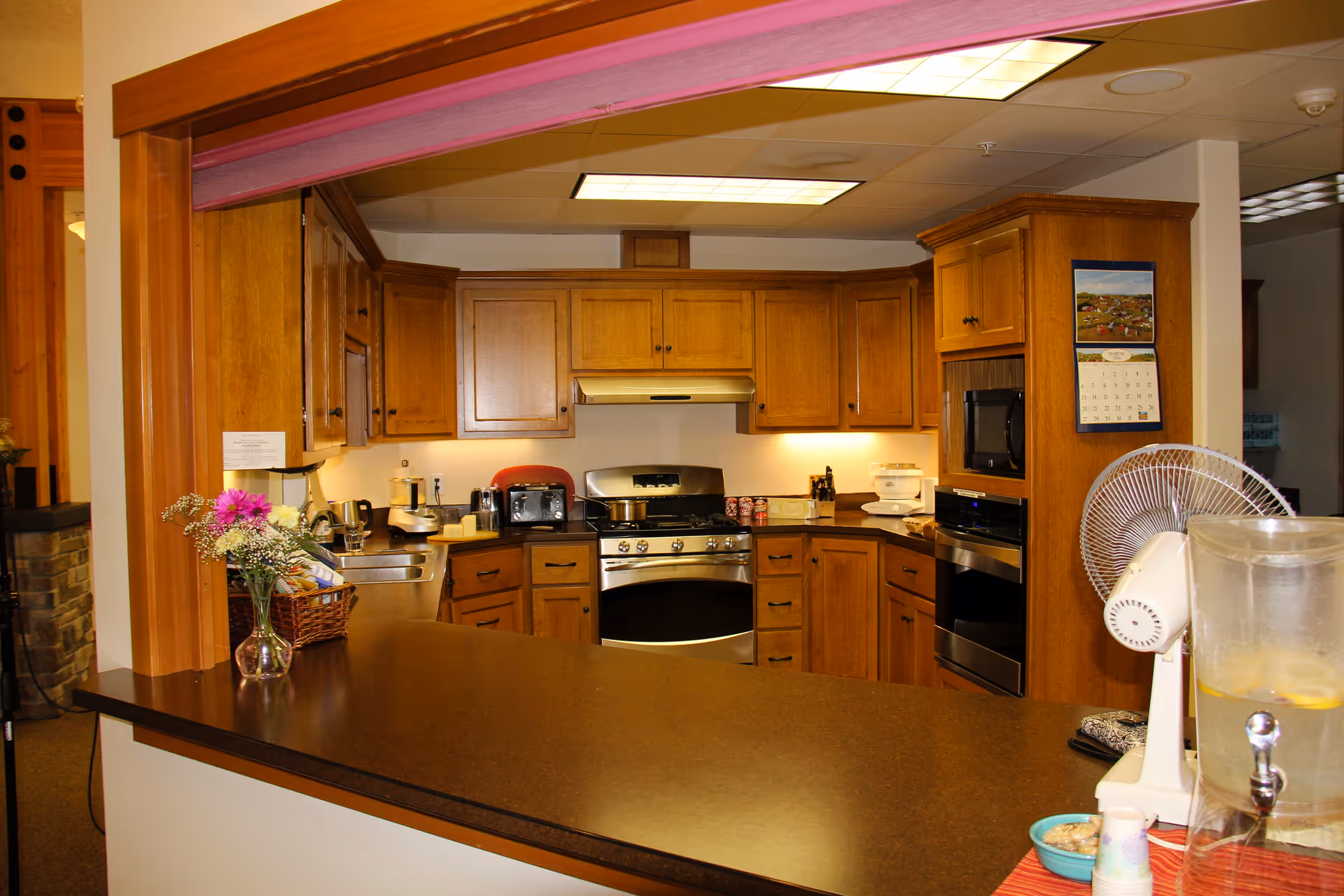 Communal kitchen area with wooden cabinets, a stainless steel stove and oven, a long front counter, and various small appliances.