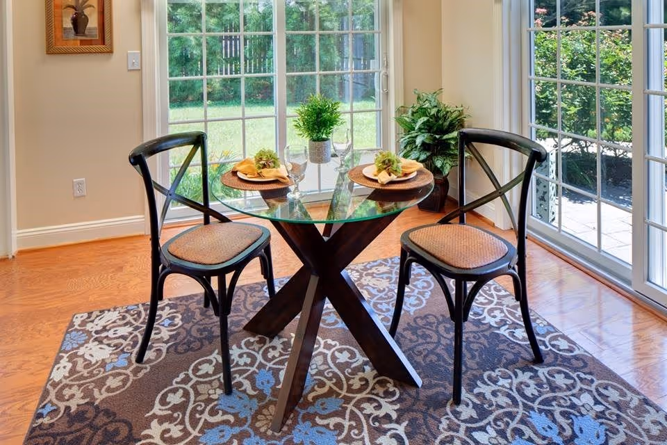 A small dining area with a round glass table and two black wooden chairs with woven seats. The table is set with two place settings, each with a napkin and glassware. The area is on a patterned rug with floral designs in blue, beige, and brown. Large sliding glass doors provide a view of a green outdoor garden space.