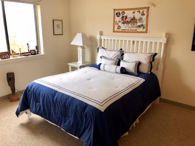 A tidy bedroom with a white wooden bed dressed in blue and white linens, a nightstand with a lamp, a window, and wall art.