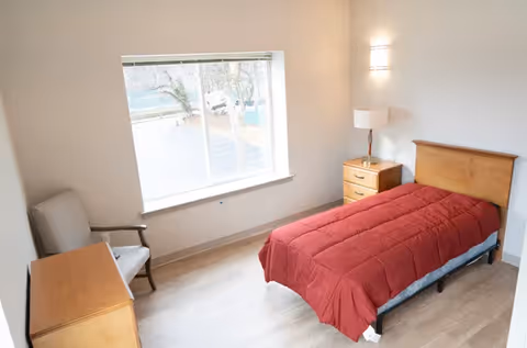 A simple, clean bedroom with a single bed covered with a red comforter, a wooden nightstand with a lamp, a wooden dresser, and a cushioned chair near a large window letting in natural light.