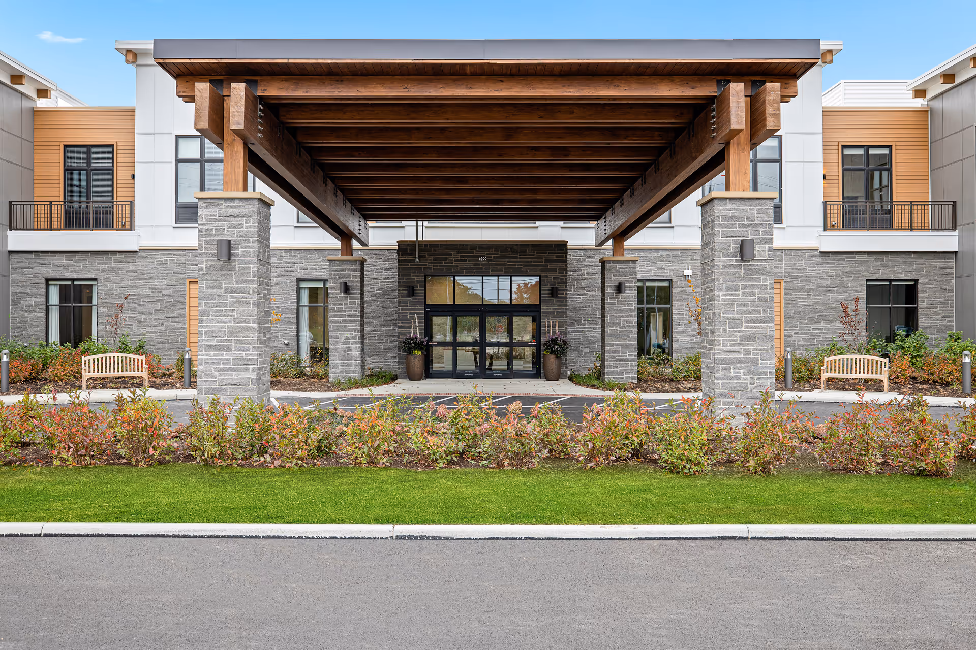 The main entrance of a senior living building with a large wooden porte-cochere supported by stone pillars, benches, and landscaped greenery.