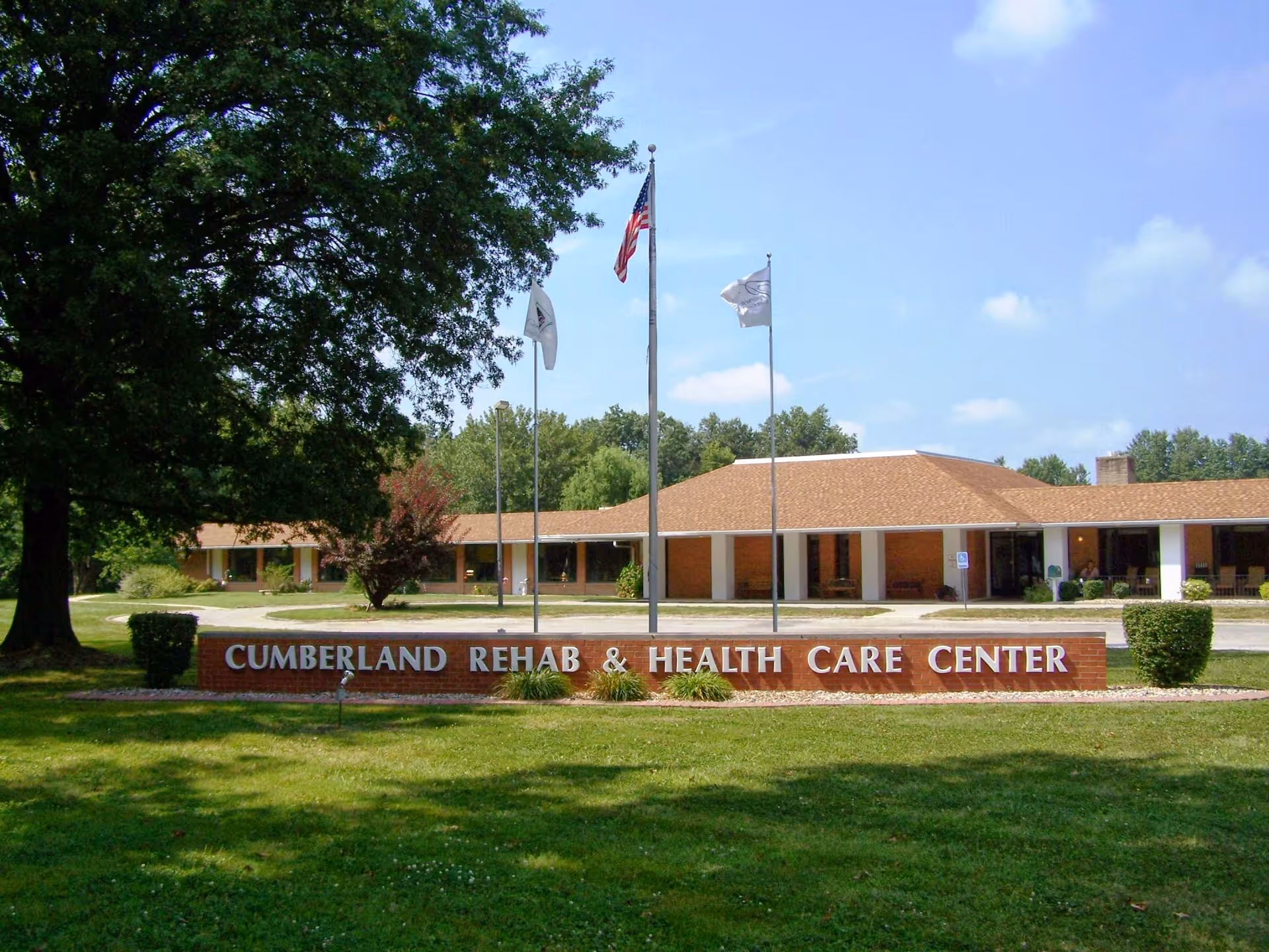 Front exterior view of Cumberland Rehab & Health Care Center building with a large sign in front, three flagpoles with flags, and surrounding trees and grass under a blue sky.