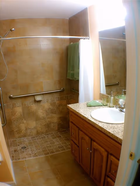 A bathroom with beige tiled walls and floor, featuring a walk-in shower with a handheld showerhead and a horizontal grab bar. There is a white shower curtain partially drawn, a wooden vanity with a granite countertop, a white sink, a mirror above the sink, and green towels hanging on a towel rack.