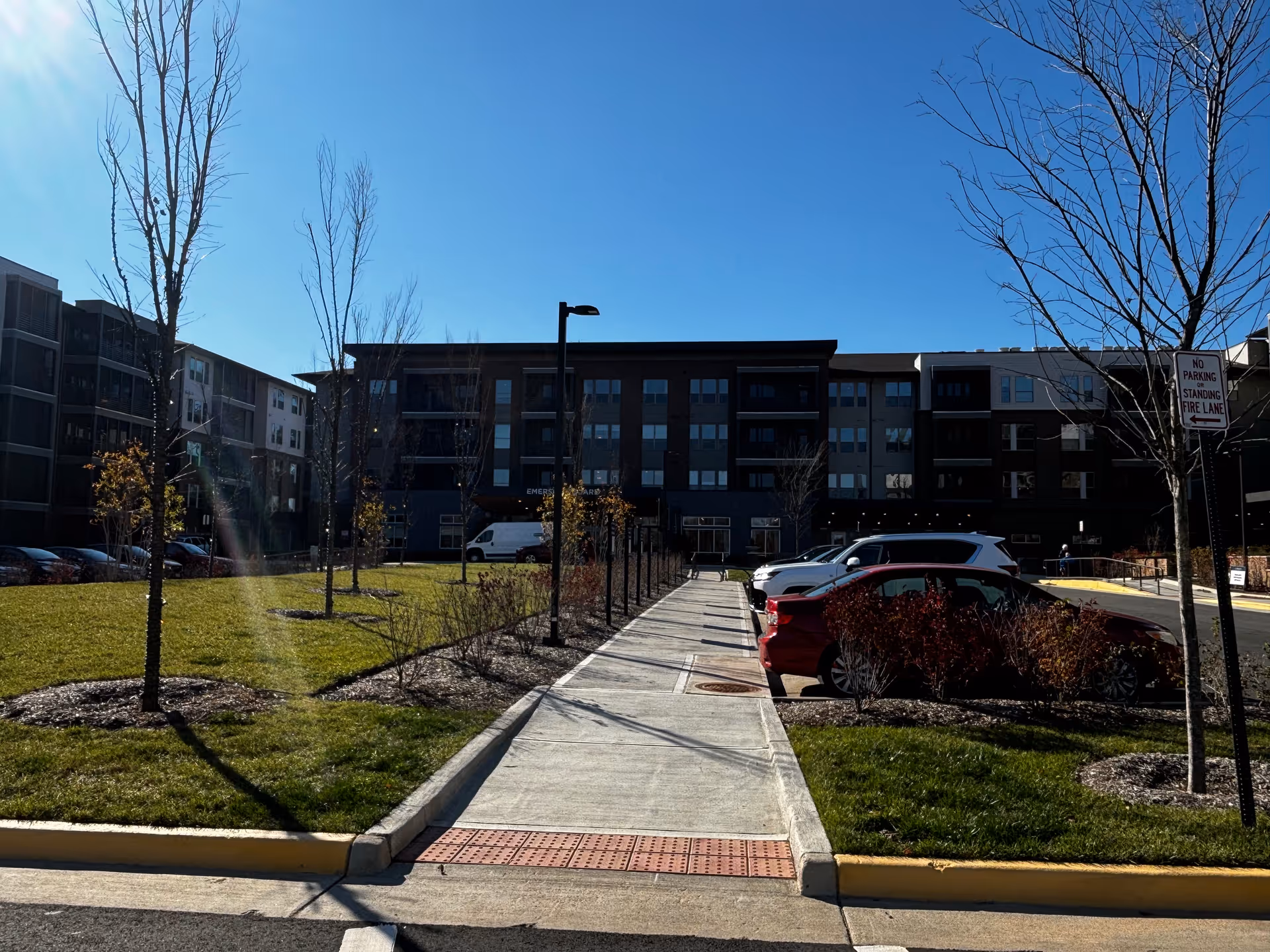 Sidewalk leading to the front entrance of a multi-story senior living building with parked cars and landscaped lawns under a clear blue sky.