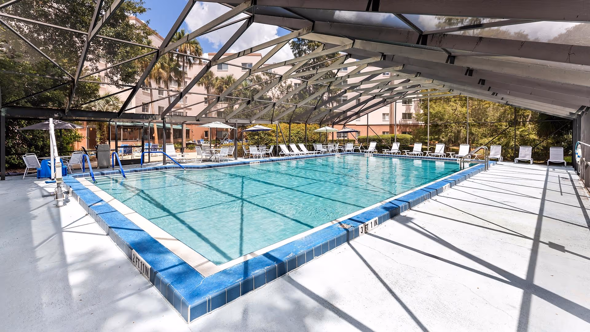 Indoor swimming pool with clear blue water surrounded by white lounge chairs and tables with umbrellas. The pool area is enclosed by a metal and glass structure allowing natural light to enter. Trees and a building are visible outside the enclosure.