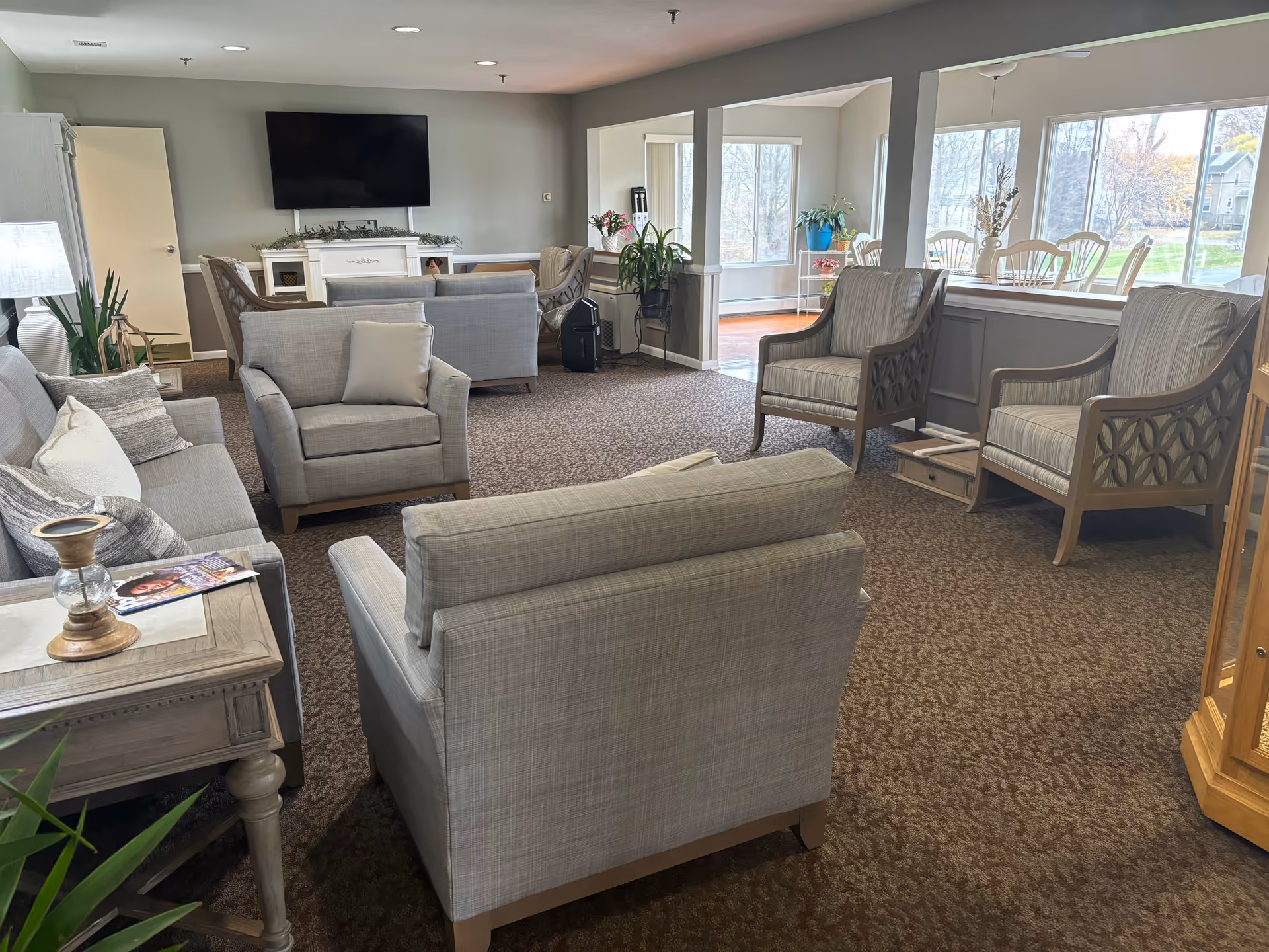 A cozy living room area in an assisted living facility with several upholstered chairs and sofas arranged around a carpeted floor. A flat-screen TV is mounted on the wall above a white fireplace mantel. Large windows let in natural light, and there are plants and decorative items placed around the room.