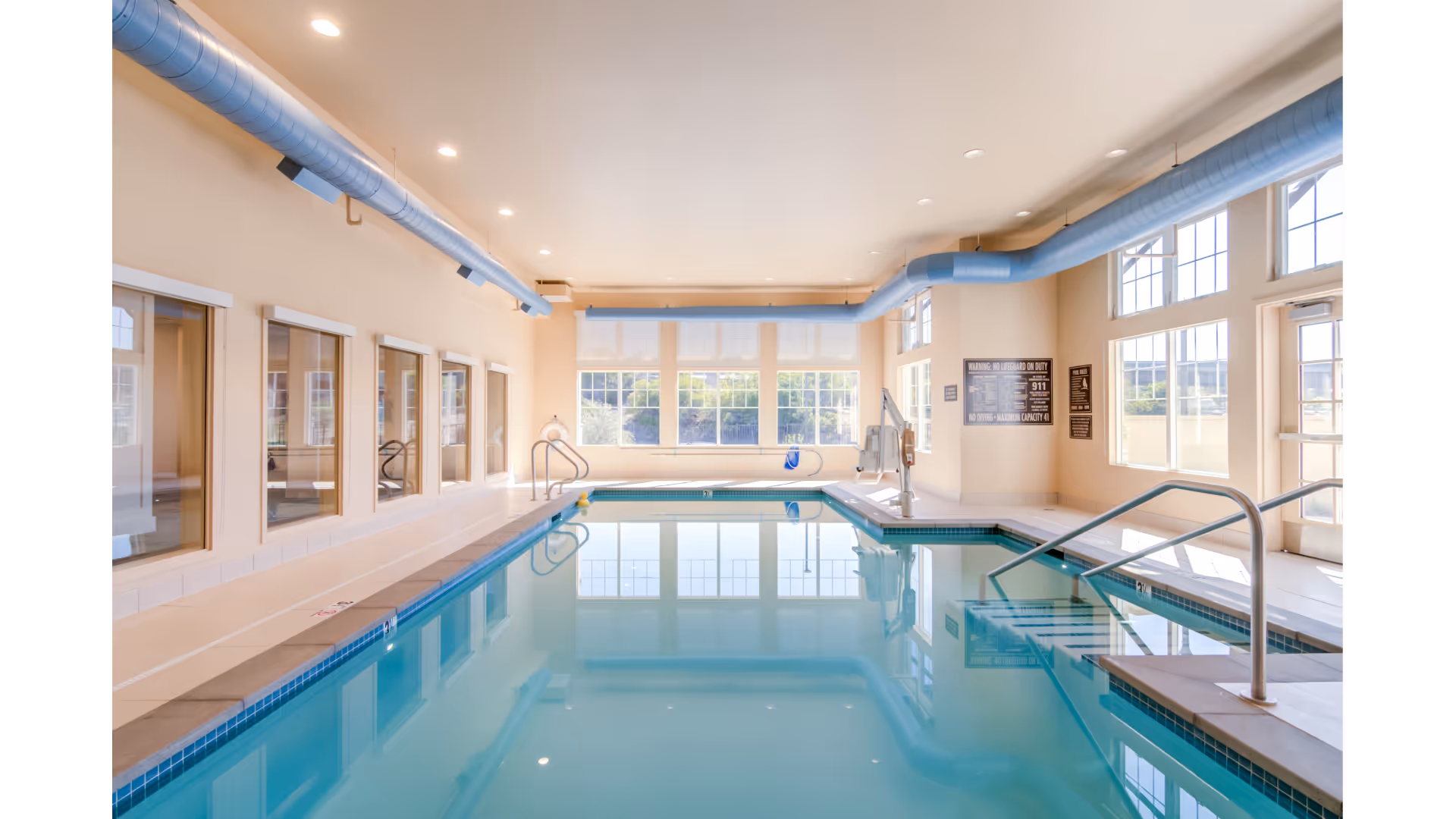 Indoor swimming pool with clear blue water, surrounded by beige tiled flooring and large windows letting in natural light. Blue ventilation ducts run along the ceiling. Pool safety signs are visible on the far wall, along with a pool lift for accessibility.