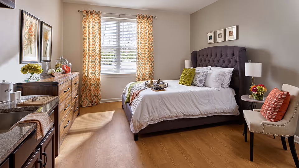 A bright and cozy bedroom with a large window covered by patterned orange and white curtains. The room features a dark upholstered bed with white bedding and colorful pillows, a wooden dresser with decorative items, a small round side table with a lamp and flowers, and a beige chair with an orange cushion. The floor is wooden, and the walls are painted light gray with framed artwork above the bed.