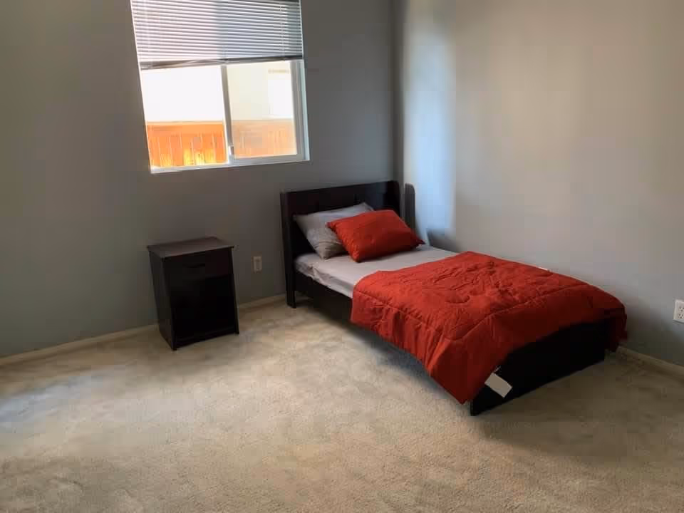 A simple bedroom with a single bed dressed in red bedding, a small dark nightstand, a window with blinds, and light gray walls.
