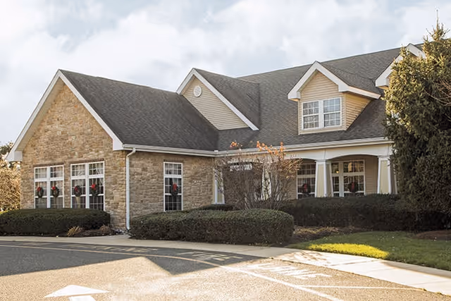 Exterior view of a single-story building with stone and beige siding, multiple windows decorated with wreaths, a covered porch, and surrounding bushes and trees under a partly cloudy sky.