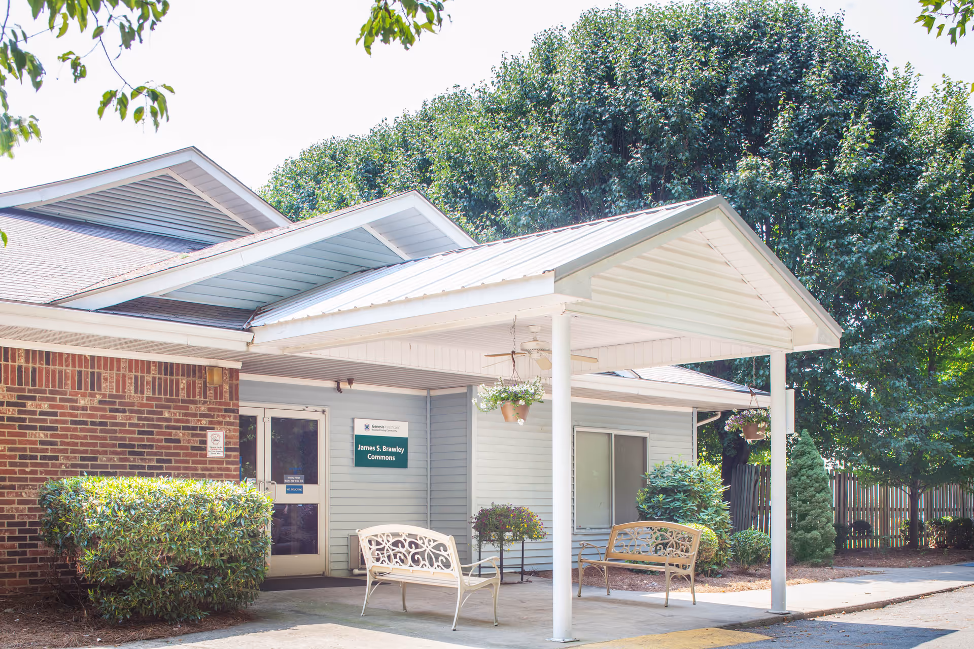 Exterior view of a senior living facility entrance with a covered porch supported by white columns. There are two metal benches under the porch, hanging flower pots, and greenery including bushes and trees surrounding the building. A sign next to the door reads 'James S. Brawley Commons.'