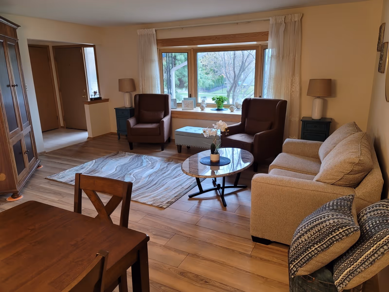 A cozy living room with a beige sofa, two brown armchairs, a round coffee table with a small plant, a patterned area rug, and a large window with sheer curtains letting in natural light. There are two side tables with lamps and decorative items on the window sill. A wooden dining table with chairs is partially visible in the foreground.