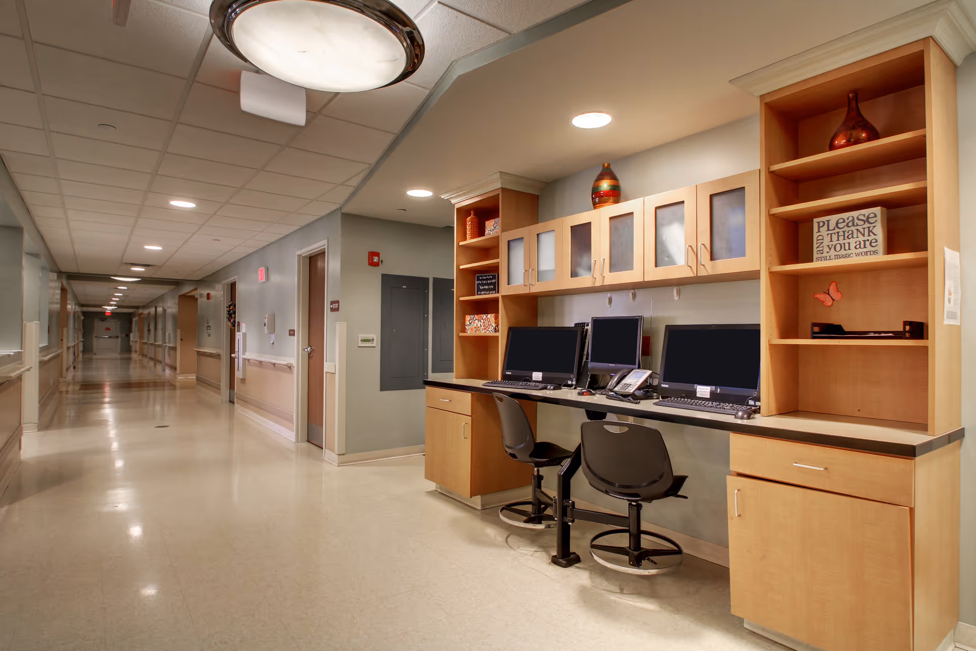 A long, clean hallway in a senior care facility with beige walls and a light-colored floor. On the right side, there is a built-in wooden desk area with three computer monitors, two office chairs, and overhead cabinets. The hallway is well-lit with ceiling lights and has handrails along the walls.