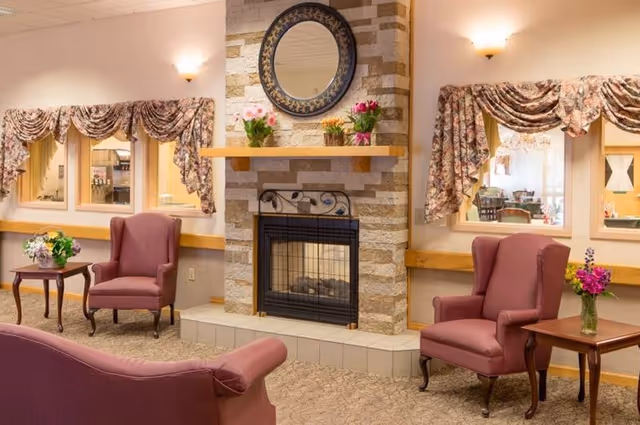 A cozy senior living common area with a stone fireplace in the center, decorated with a round mirror and flower vases on the mantel. There are two mauve wingback chairs on either side of the fireplace, each next to a wooden side table with flower arrangements. The room has floral valance curtains over windows that look into another room.