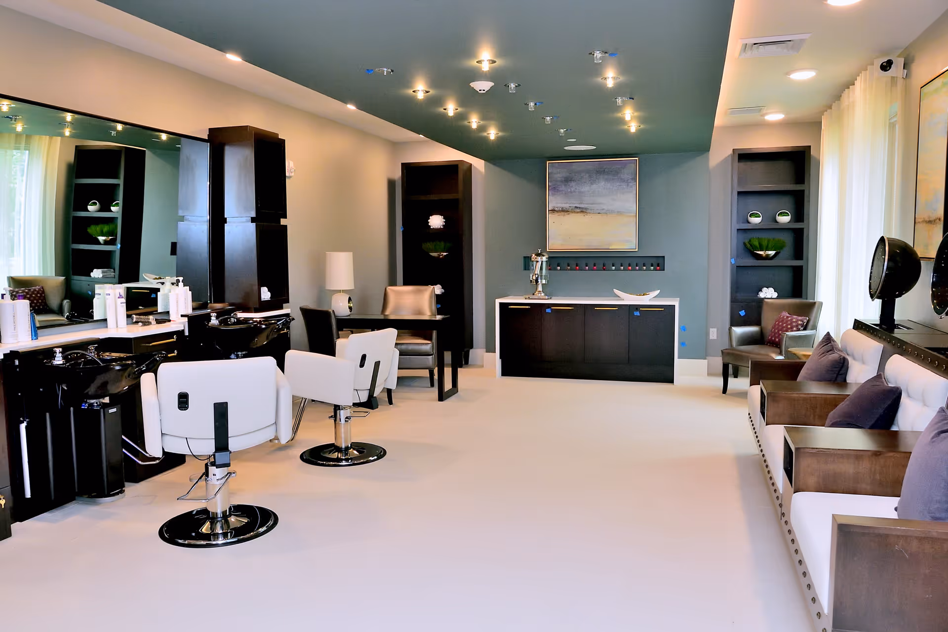 A modern salon area in a senior living facility with white styling chairs and black wash basins on the left, a large mirror behind them, and a seating area with armchairs and a sofa with purple cushions on the right. The room has soft lighting from ceiling fixtures and decorative shelves with plants and ornaments.