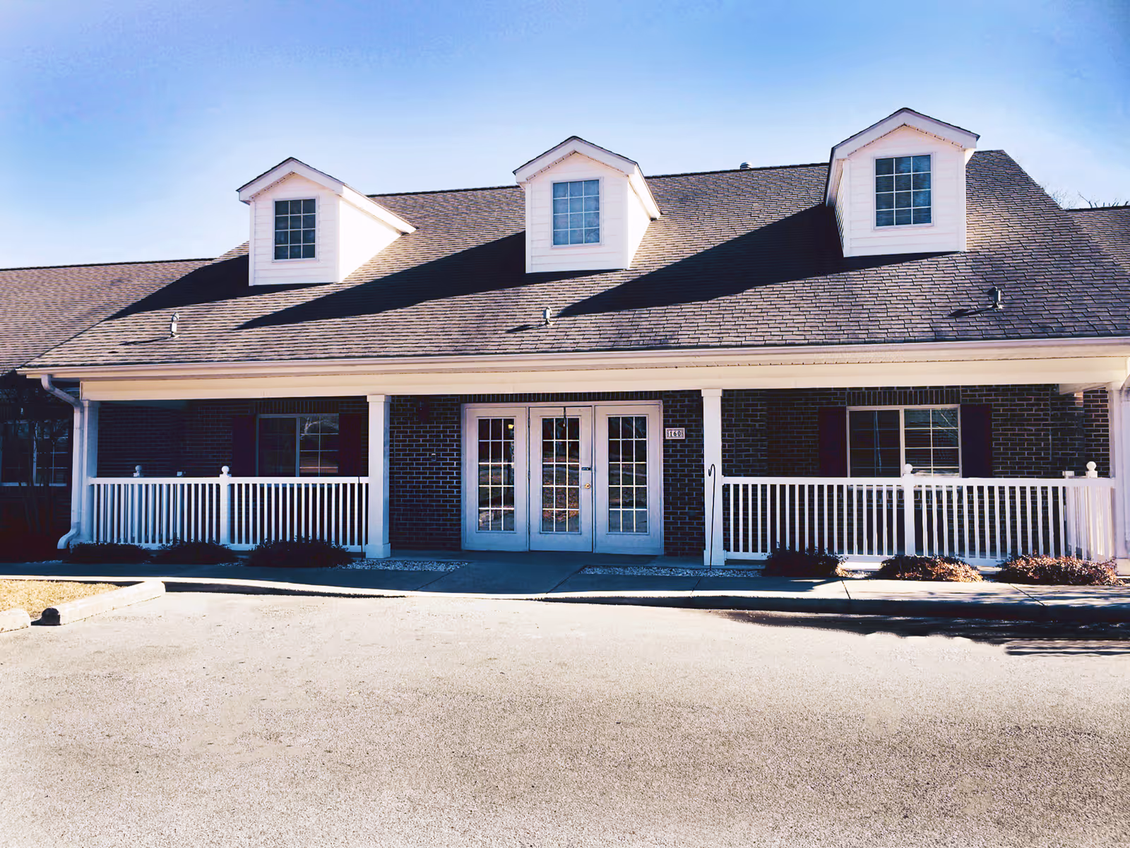 Front exterior view of a single-story brick building with a dark shingled roof featuring three dormer windows. The building has white framed double glass doors in the center, flanked by windows with dark shutters. A white railing runs along the front porch area under the roof overhang. The sky is clear and blue.