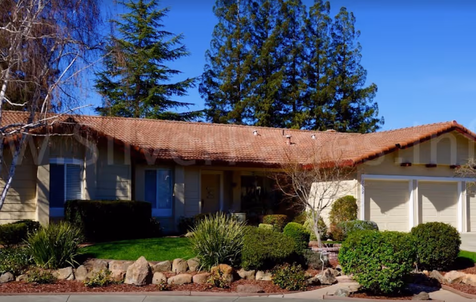 Single-story residential building with a tiled roof, beige exterior walls, and a three-car garage. The front yard is landscaped with green bushes, small trees, rocks, and a well-maintained lawn. Tall evergreen trees are visible in the background under a clear blue sky.