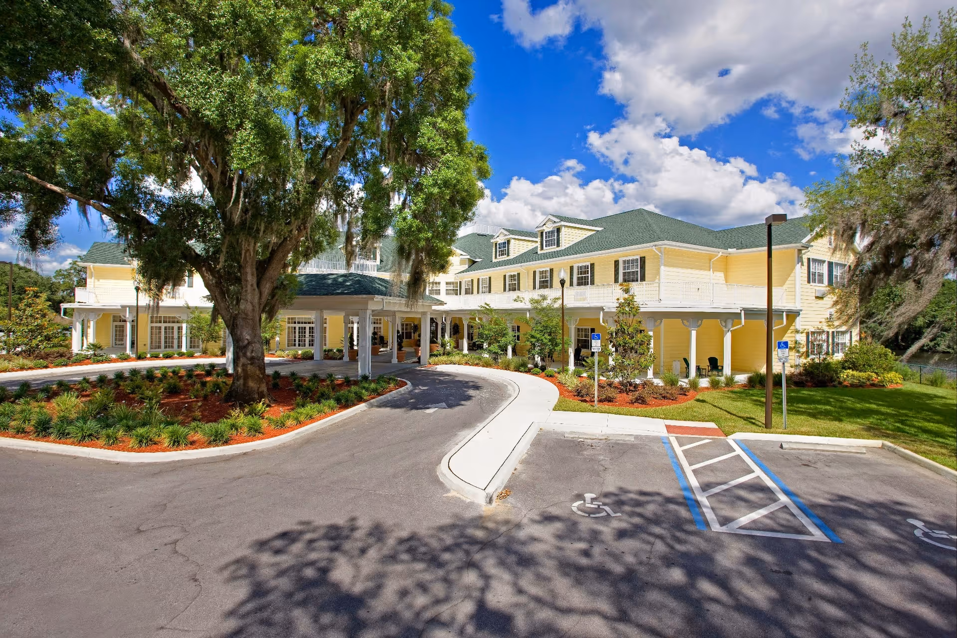 Exterior view of a large yellow senior living facility building with a green roof, surrounded by landscaped gardens and large trees under a partly cloudy blue sky. There is a circular driveway with handicap parking spaces in front of the building entrance.