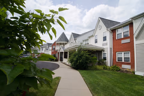 Exterior view of a senior living facility with a paved walkway, green lawn, bushes, and a multi-story building featuring white siding and red brick accents under a partly cloudy sky.