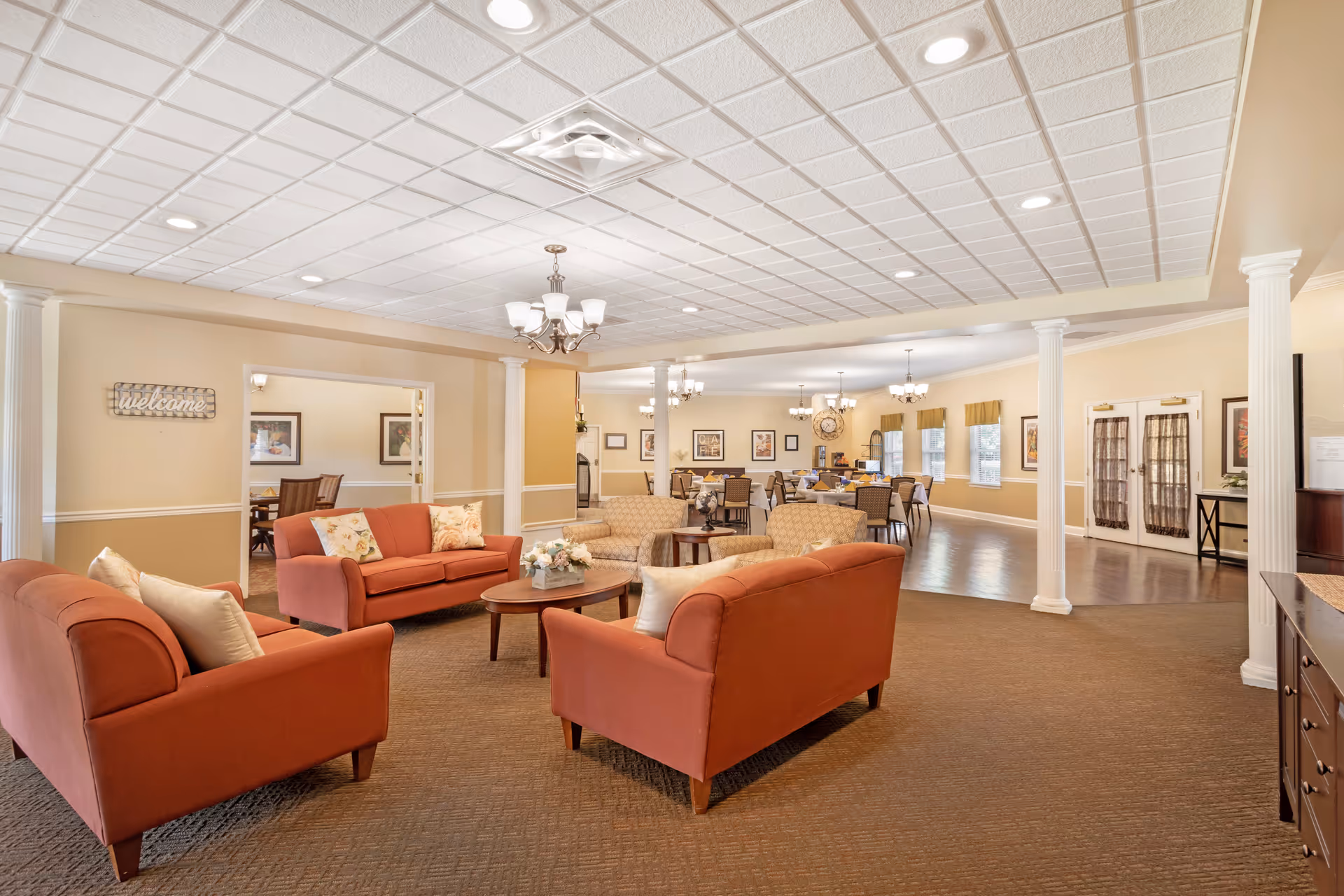 A spacious and well-lit common area in a senior living facility featuring multiple seating arrangements with orange and beige sofas and armchairs around wooden coffee tables. The room has beige walls, white columns, and a tiled ceiling with recessed lighting and chandeliers. In the background, there is a dining area with tables and chairs, framed artwork on the walls, and large windows with valances.