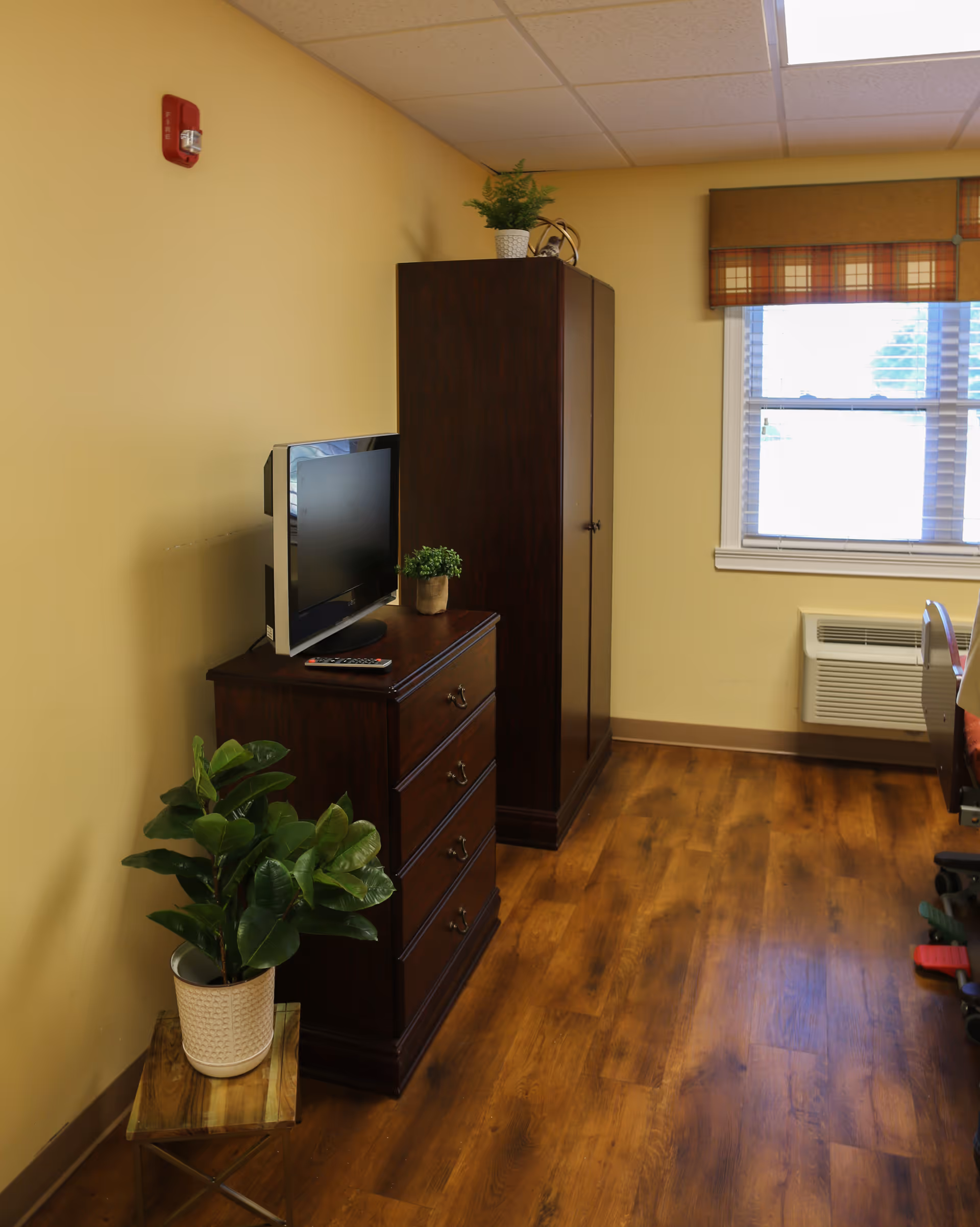 A corner of a room with wooden flooring and beige walls. There is a dark wooden dresser with a flat-screen TV and a small potted plant on top. Next to the dresser is a tall dark wooden wardrobe with a potted plant on top. A window with blinds and a plaid valance is on the right wall, and below it is a wall-mounted air conditioning unit. A larger potted plant sits on a small wooden stand on the floor near the dresser.