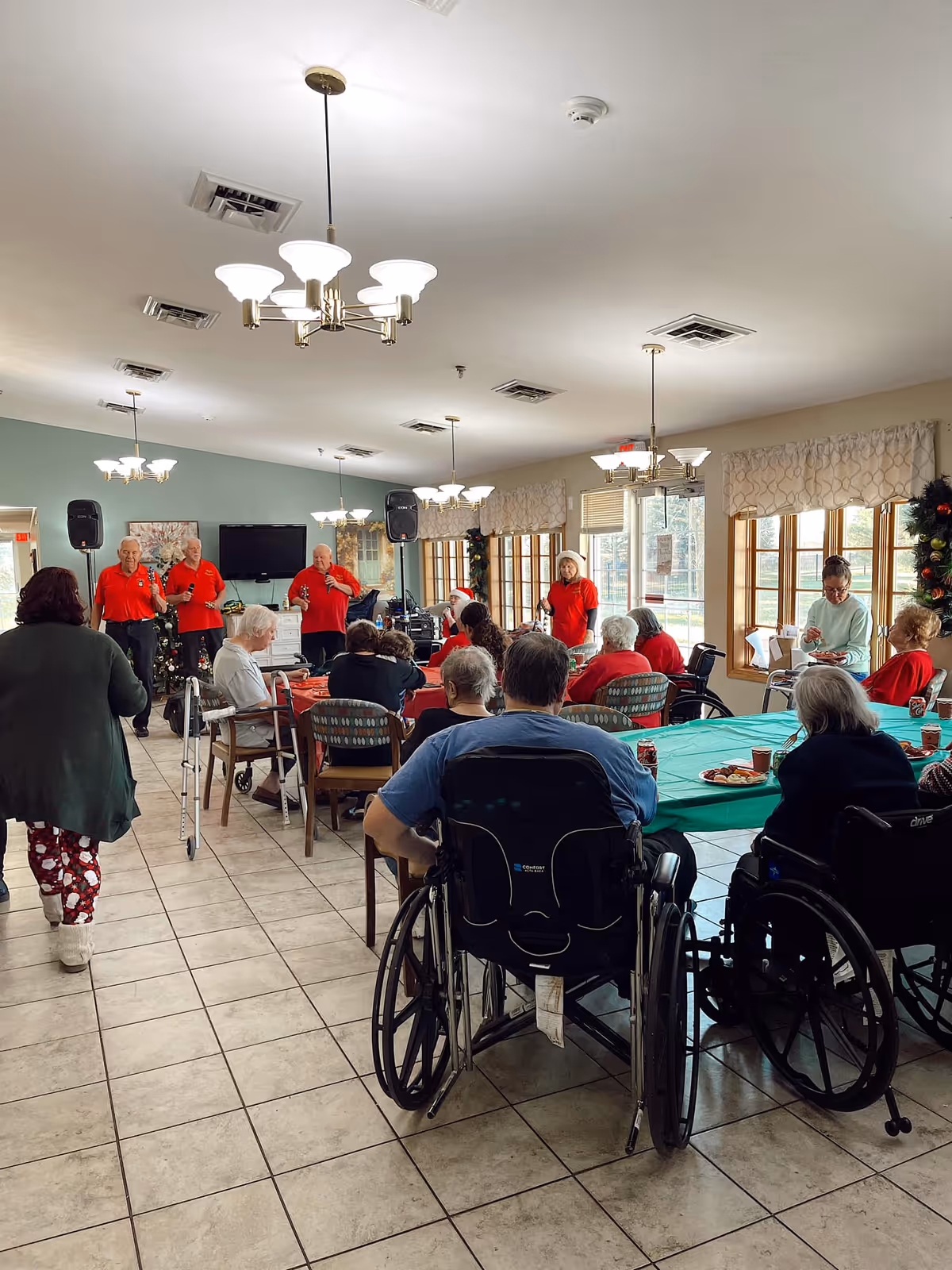 Residents and staff gathered in a senior living dining room for a group event, with tables, wheelchairs, and performers at the front.