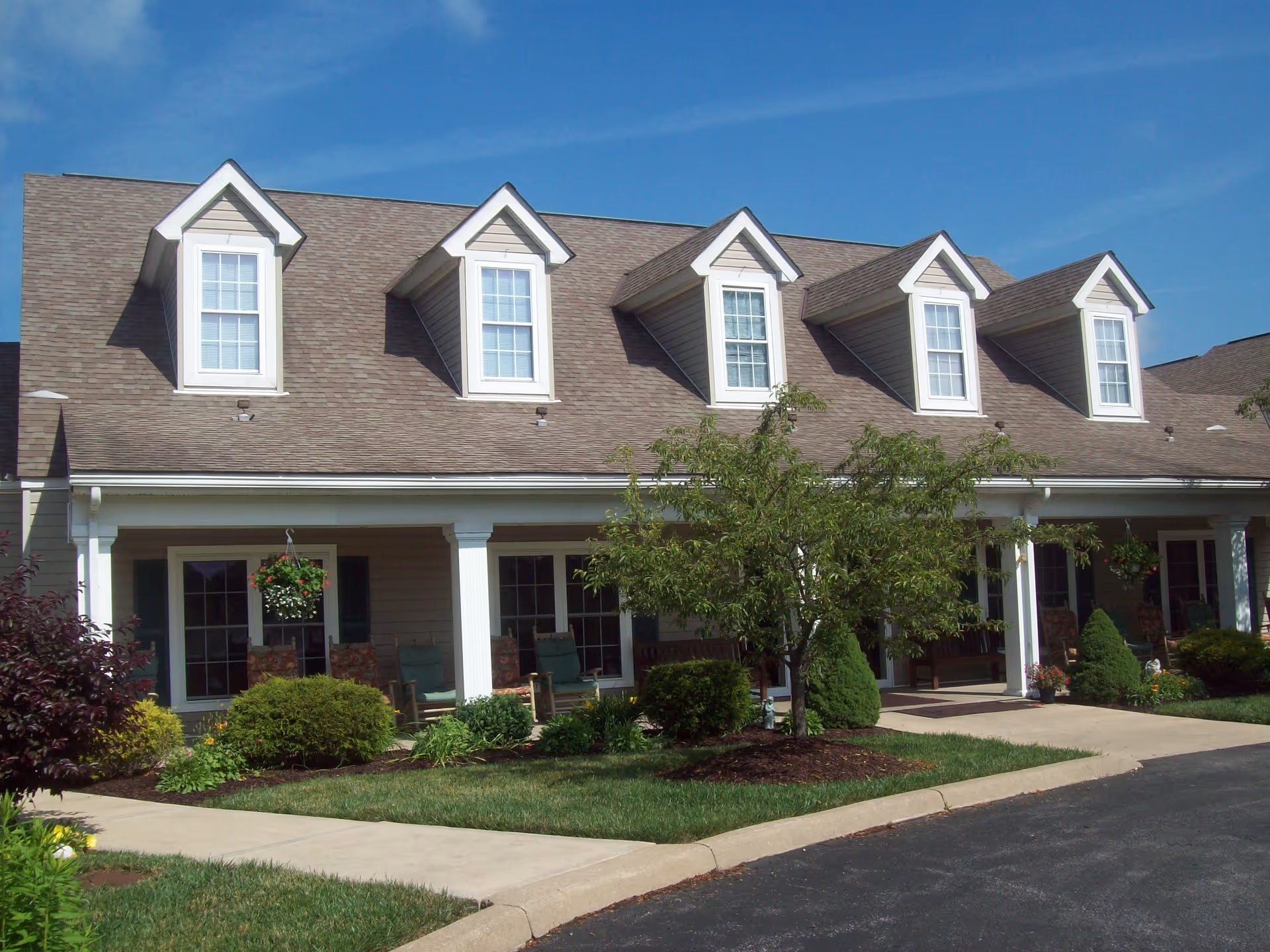 Front exterior of a building with a covered porch, dormer windows, landscaping and a curved driveway.