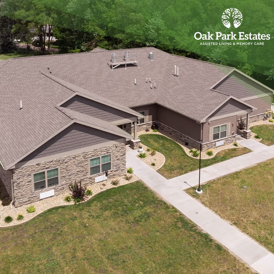Aerial view of a single-story assisted living facility building with stone and siding exterior, surrounded by green lawn and trees. Concrete walkways lead to the entrances. The Oak Park Estates Assisted Living and Memory Care logo is visible in the top right corner.