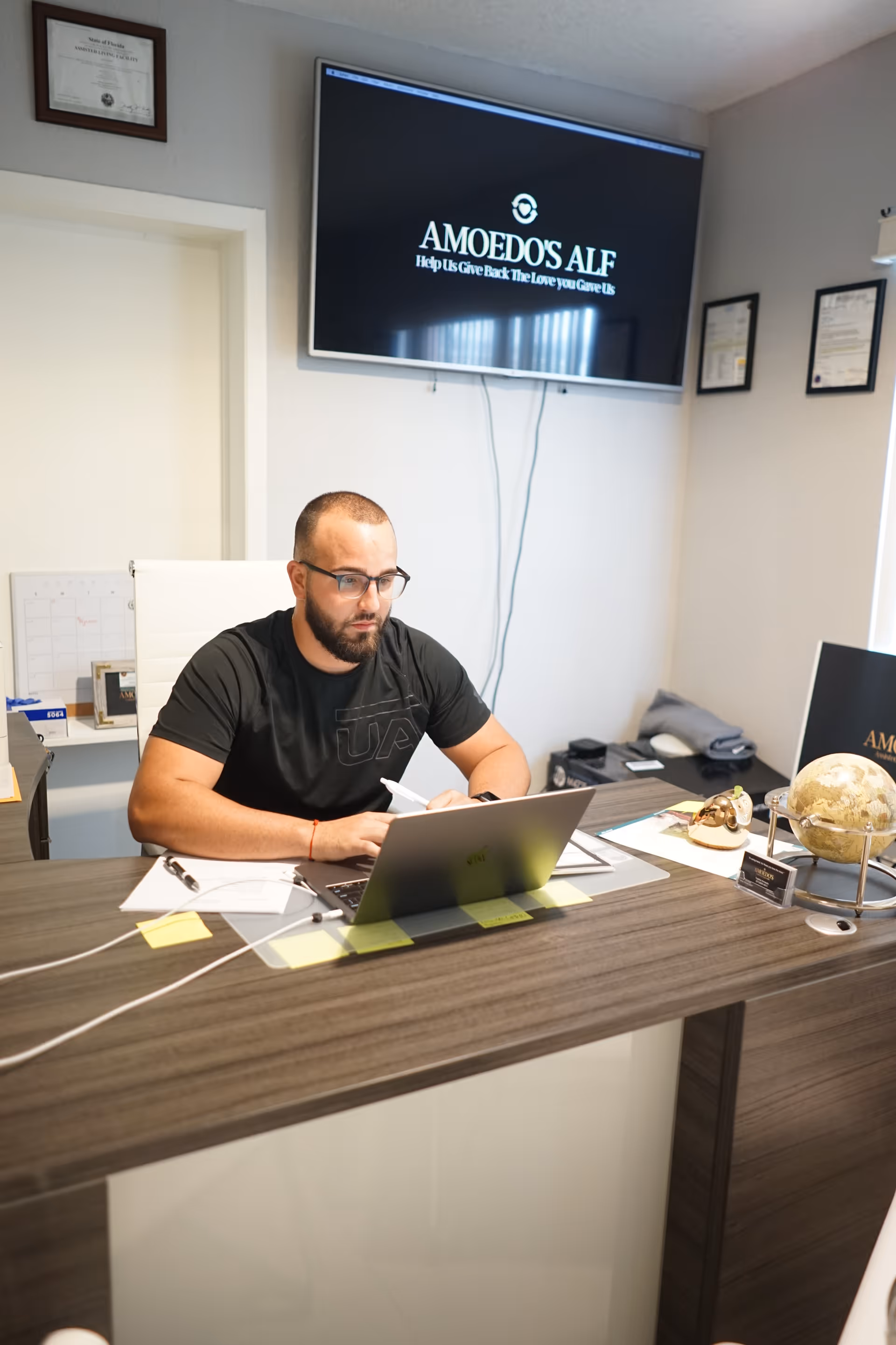 A man with glasses and a beard is sitting at a desk working on a laptop in an office. Behind him, a wall-mounted TV screen displays the text 'AMOEDO'S ALF Help Us Give Back The Love you Gave Us'. The office has framed certificates on the wall and various office supplies on the desk, including a globe and a model of a skull.