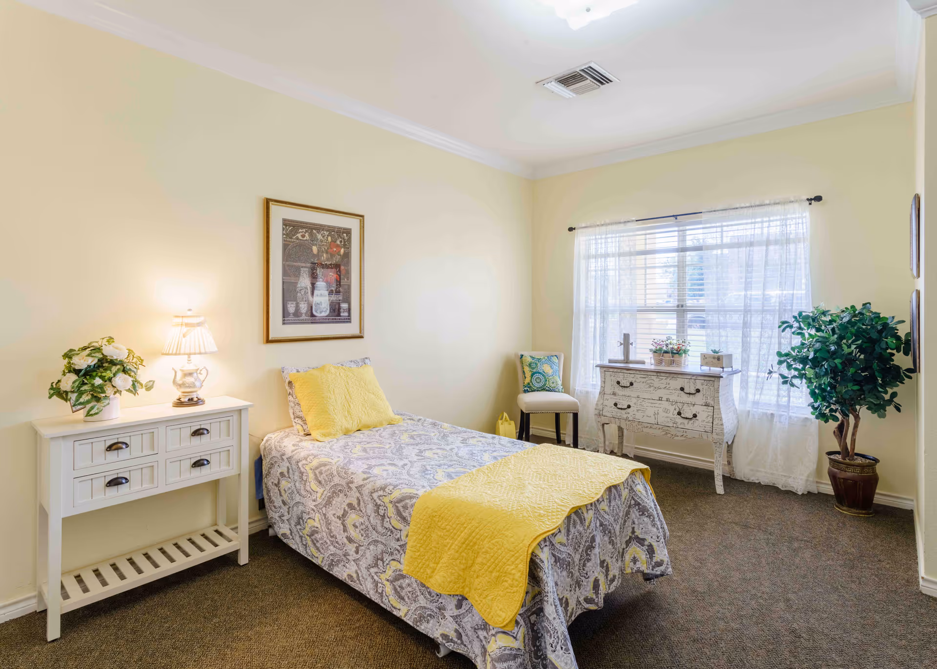 A bright and cozy bedroom with a single bed covered in a patterned bedspread with yellow accents. There is a white side table with a lamp and a flower arrangement on the left side of the bed. A framed picture hangs on the wall above the bed. On the right side of the room, there is a window with sheer white curtains, a decorative white dresser with floral and script designs, a chair with a colorful cushion, and a potted plant in the corner.