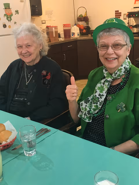 Two elderly women sitting at a table in a kitchen or dining area. One woman is wearing a green hat and scarf with shamrock patterns, giving a thumbs up and smiling. The other woman is smiling and wearing a dark outfit. On the table are glasses of water, utensils, and a basket with bread rolls. The background shows kitchen cabinets, a refrigerator with shamrock decorations, and various kitchen items.