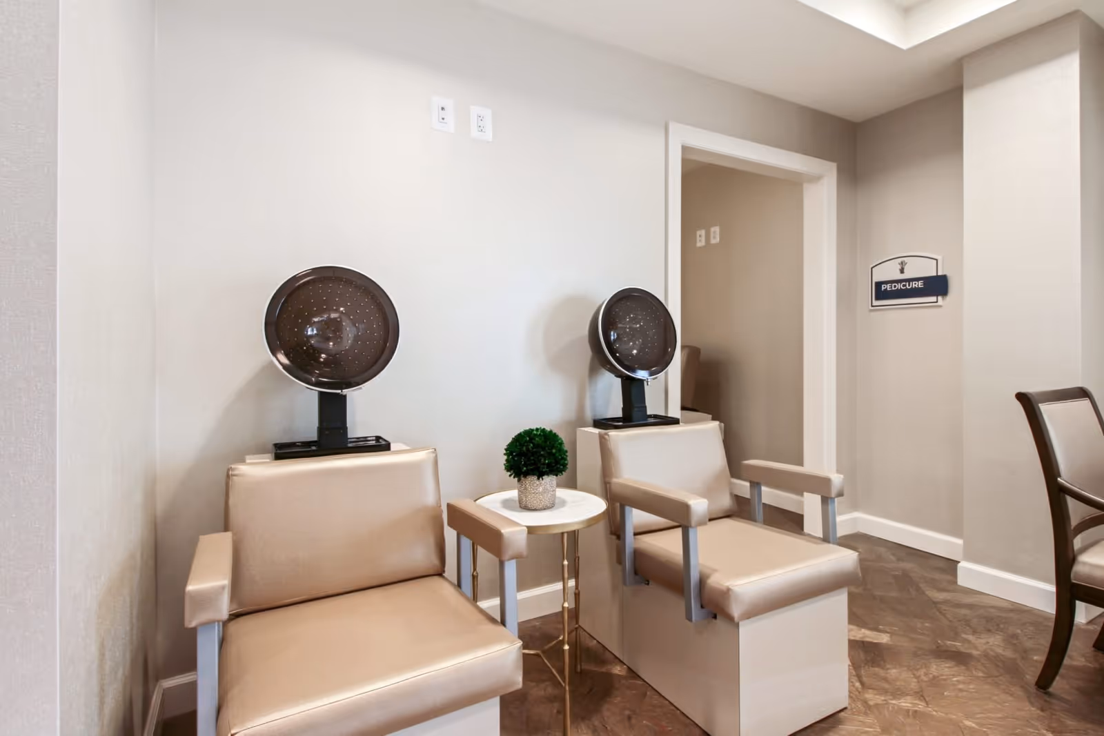 Two beige pedicure chairs with overhead dryers and a small table in a spa-like interior with a 'PEDICURE' sign on the wall.