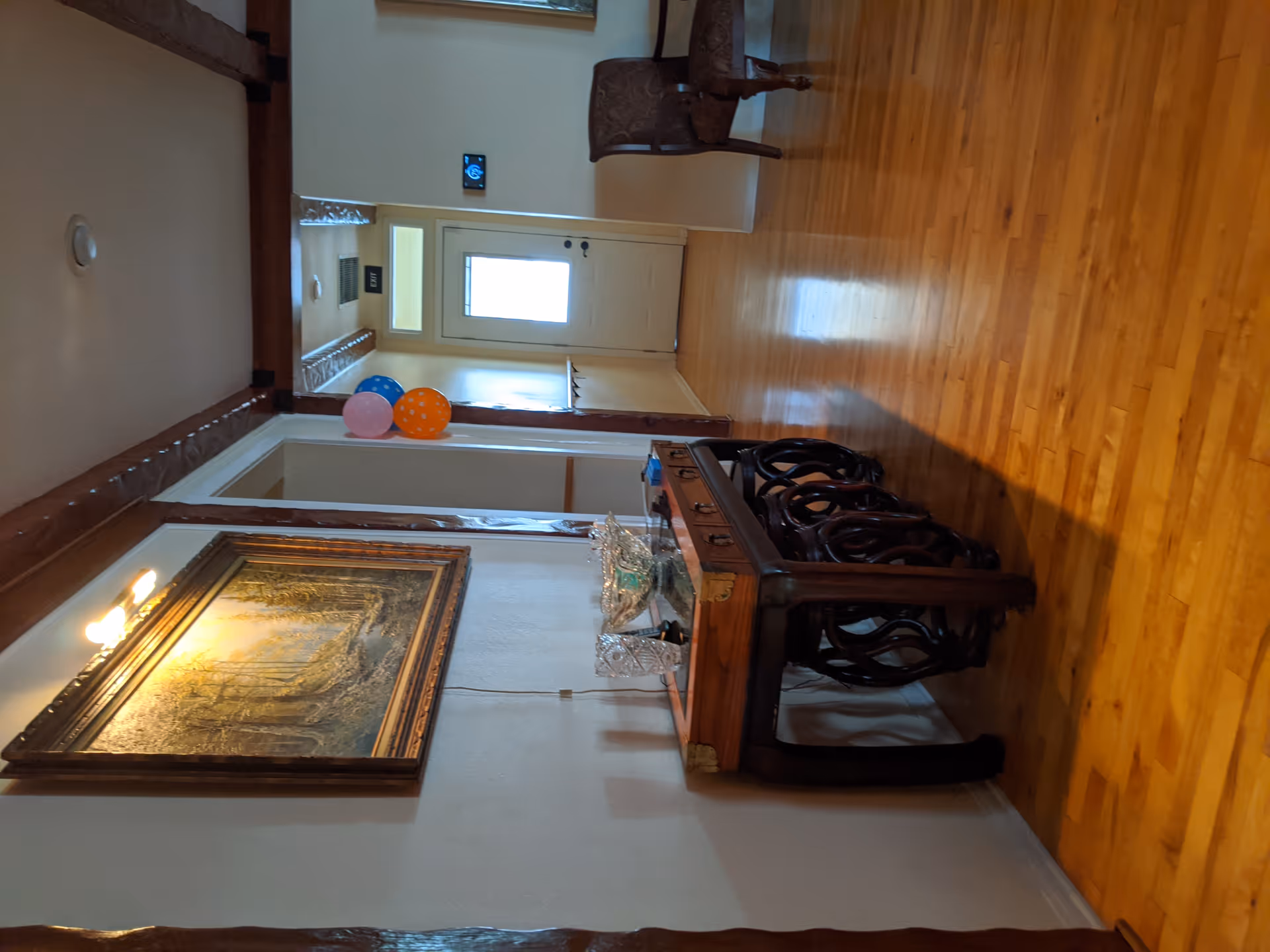 Interior view of a hallway in a senior living facility with polished wooden floors, a wooden table with decorative items, a framed painting on the wall, and a door at the end of the hallway. There are three balloons (blue, pink, and orange) near the door and a wooden chair against the wall.