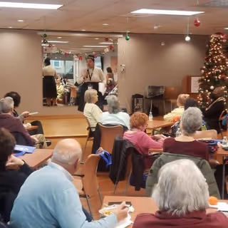 A group of elderly people seated at tables in a common room, facing a woman standing and speaking near a large mirror. The room is decorated with Christmas ornaments and a decorated Christmas tree is visible on the right side.