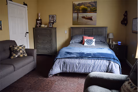 A cozy bedroom with a bed covered in patterned bedding and a blue throw blanket, flanked by a nightstand with a lamp and a digital clock. There is a gray armchair and a gray sofa with patterned pillows. A tall dresser with decorative items and framed pictures stands against the wall. A painting of a boat on a lake hangs above the bed. The walls are painted beige and the floor is carpeted.