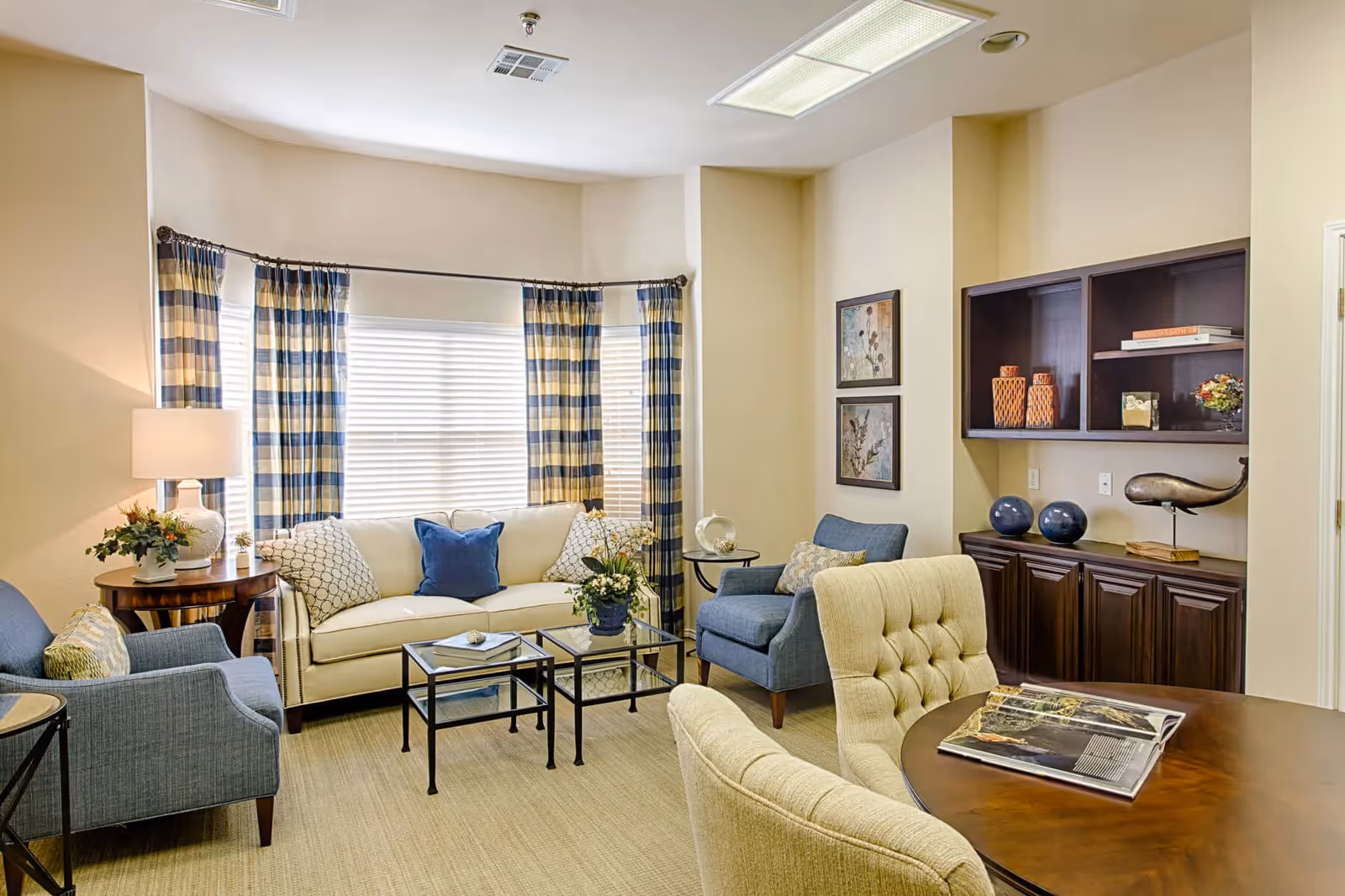 Bright furnished living room with a sofa, armchairs, coffee tables, patterned curtains and built-in shelving.