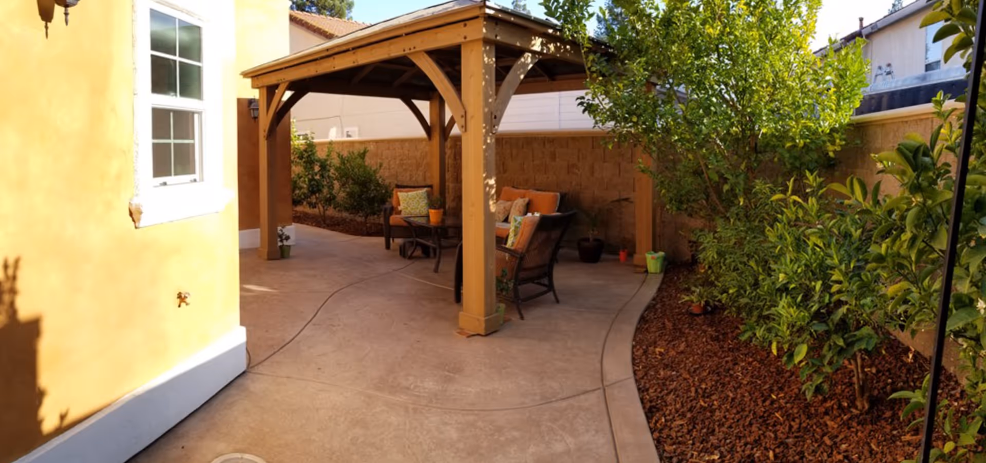 Covered patio with a wooden pergola, outdoor seating, and a paved walkway flanked by landscaping.
